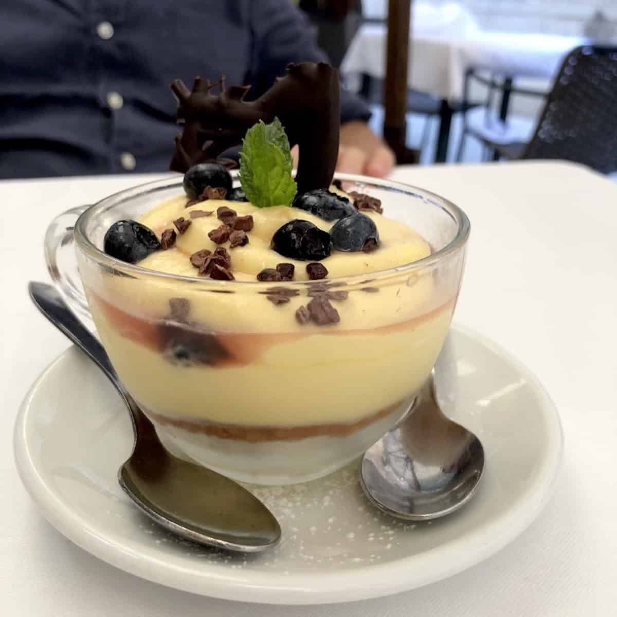 Blueberry tiramisu from Antiche Carampane fish restaurant in Venice served in a glass mug with blueberry sauce layers, ladyfingers, fresh blueberries, mint, and chocolate garnish.
