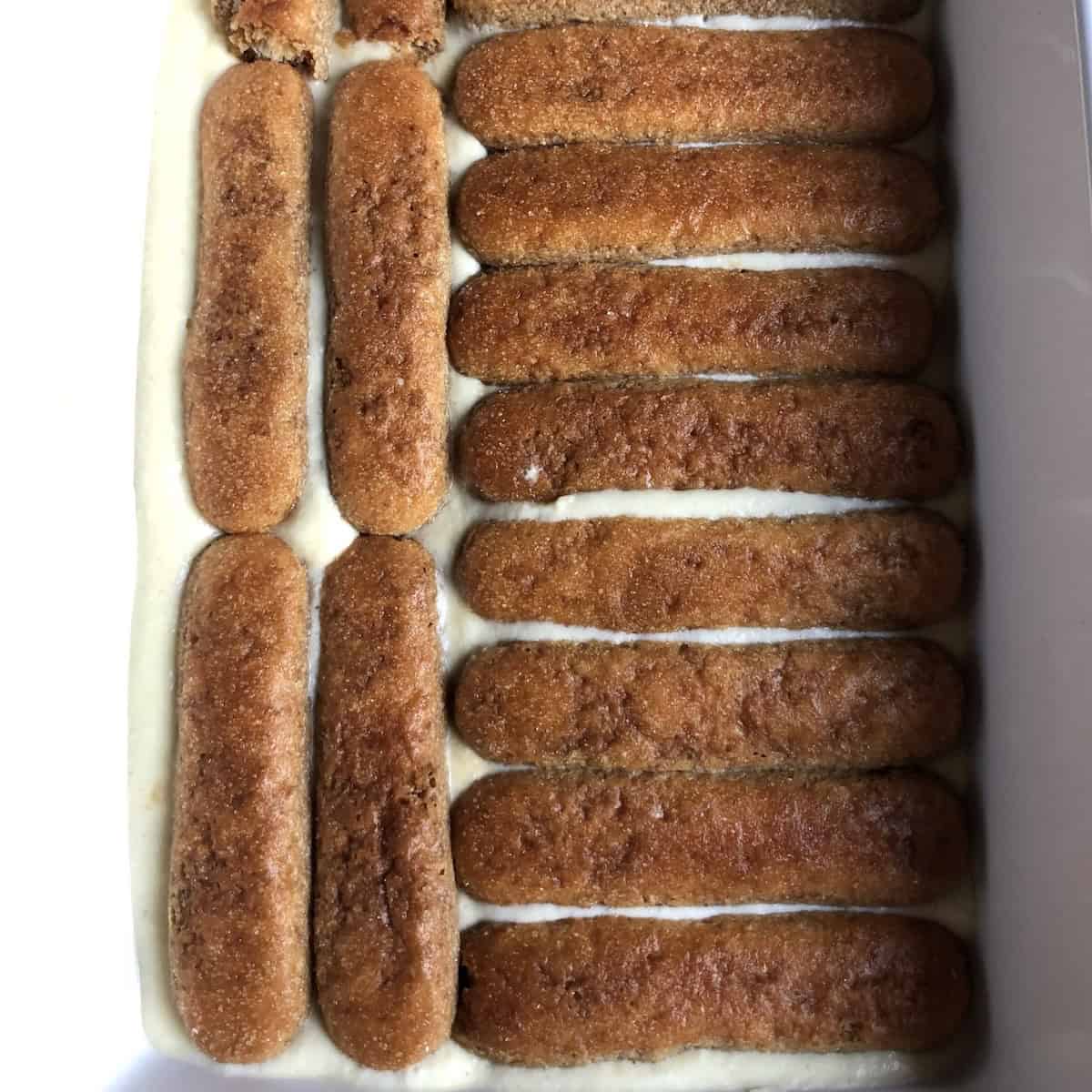 Espresso soaked ladyfingers layered on top of tiramisu cream in a 9 by 13 white ceramic casserole dish.

