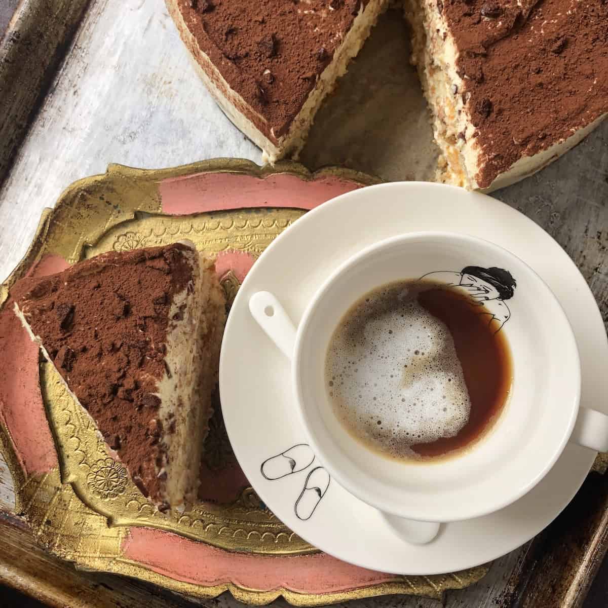 Three-layer slice of authentic Italian tiramisu on a tray with a 9-inch round tiramisu in the background, best tiramisu recipe with creamy mascarpone layers.