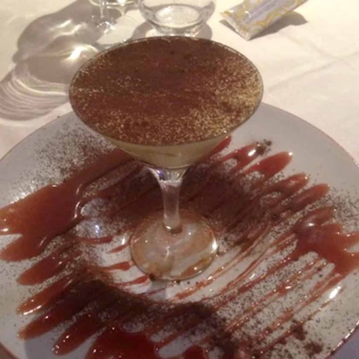 Italian restaurant tiramisu served in a martini glass dusted with cocoa powder on a plate drizzled with strawberry sauce.


