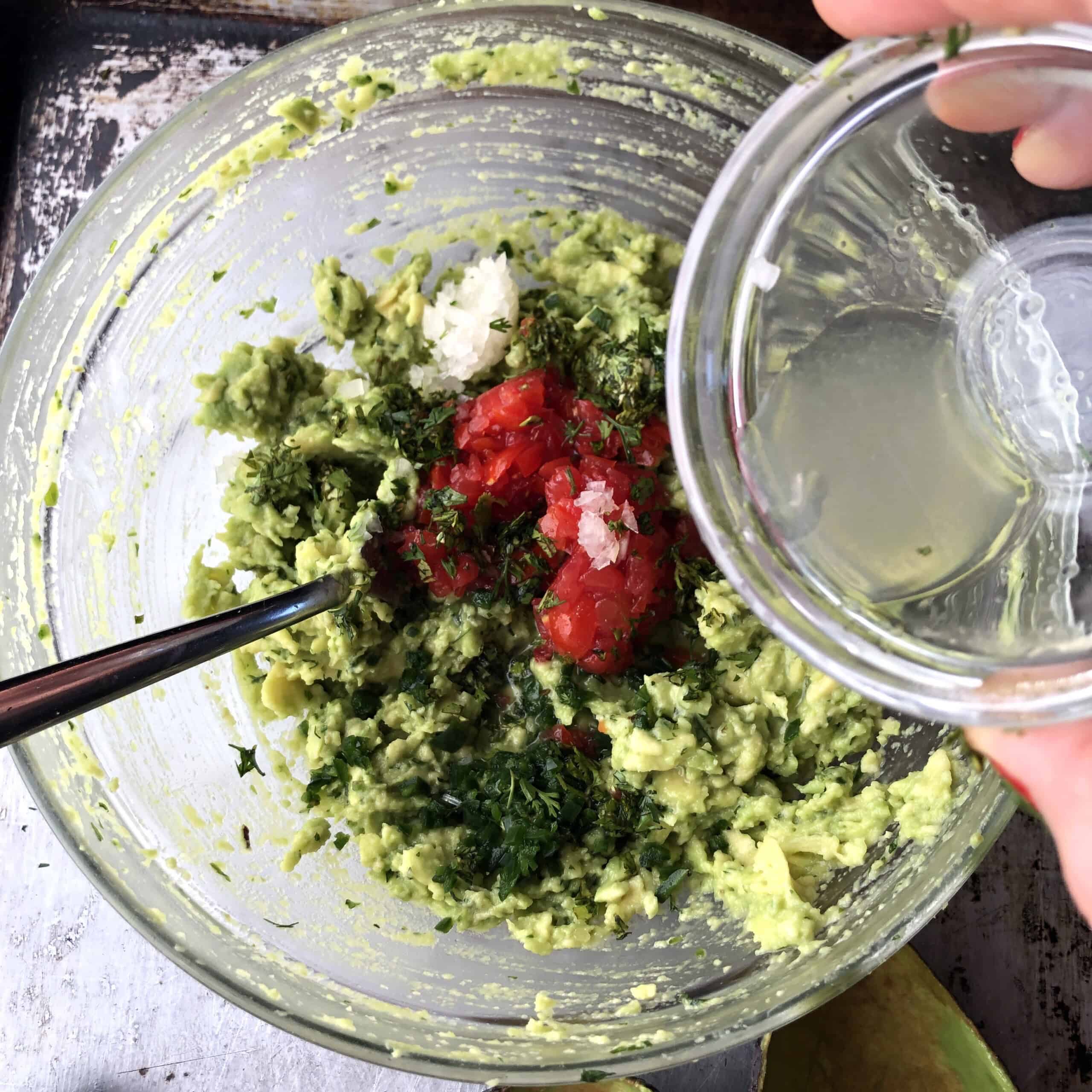 Pouring fresh lime juice over diced tomatoes and remaining aromatics added to the avocado mixture in a bowl.
