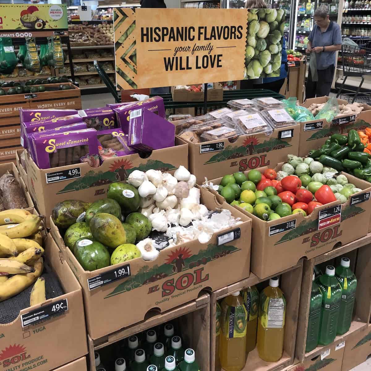 Grocery store display of Hispanic food items including avocados, onions, tomatoes, and limes for making guacamole with a sign reading "Hispanic flavors your family will love".