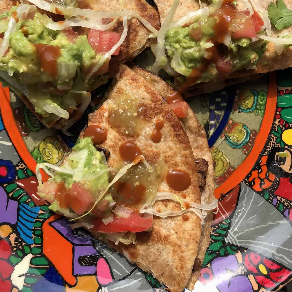 Three homemade quesadilla wedges topped with homemade guacamole, pico de gallo, hot sauce, and shredded lettuce on a plate.