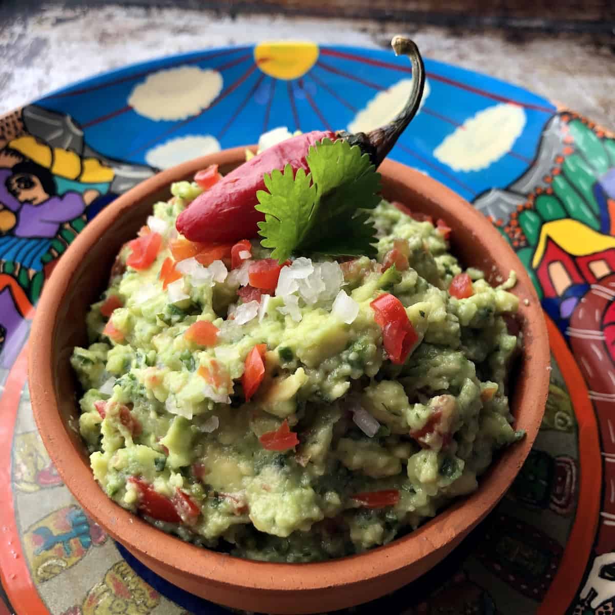 Easy guacamole recipe in a clay serving dish set on a colorful Mexican plate, simple homemade guacamole.