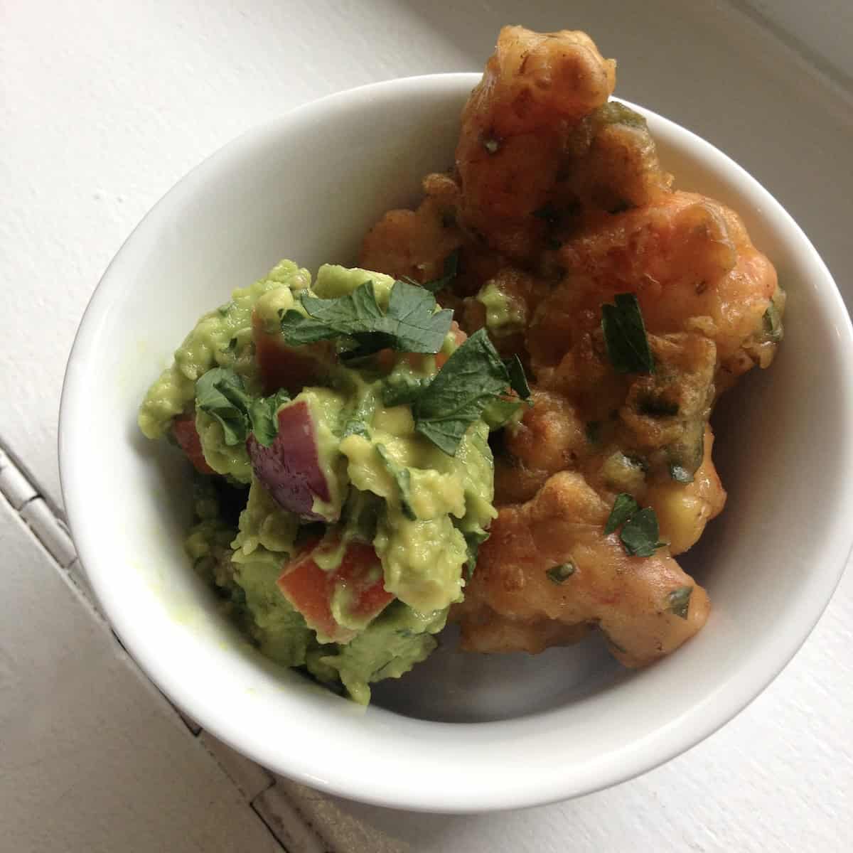 Two homemade Argentinian red shrimp and corn fritters in a ceramic dish with a scoop of guacamole with red onions.
