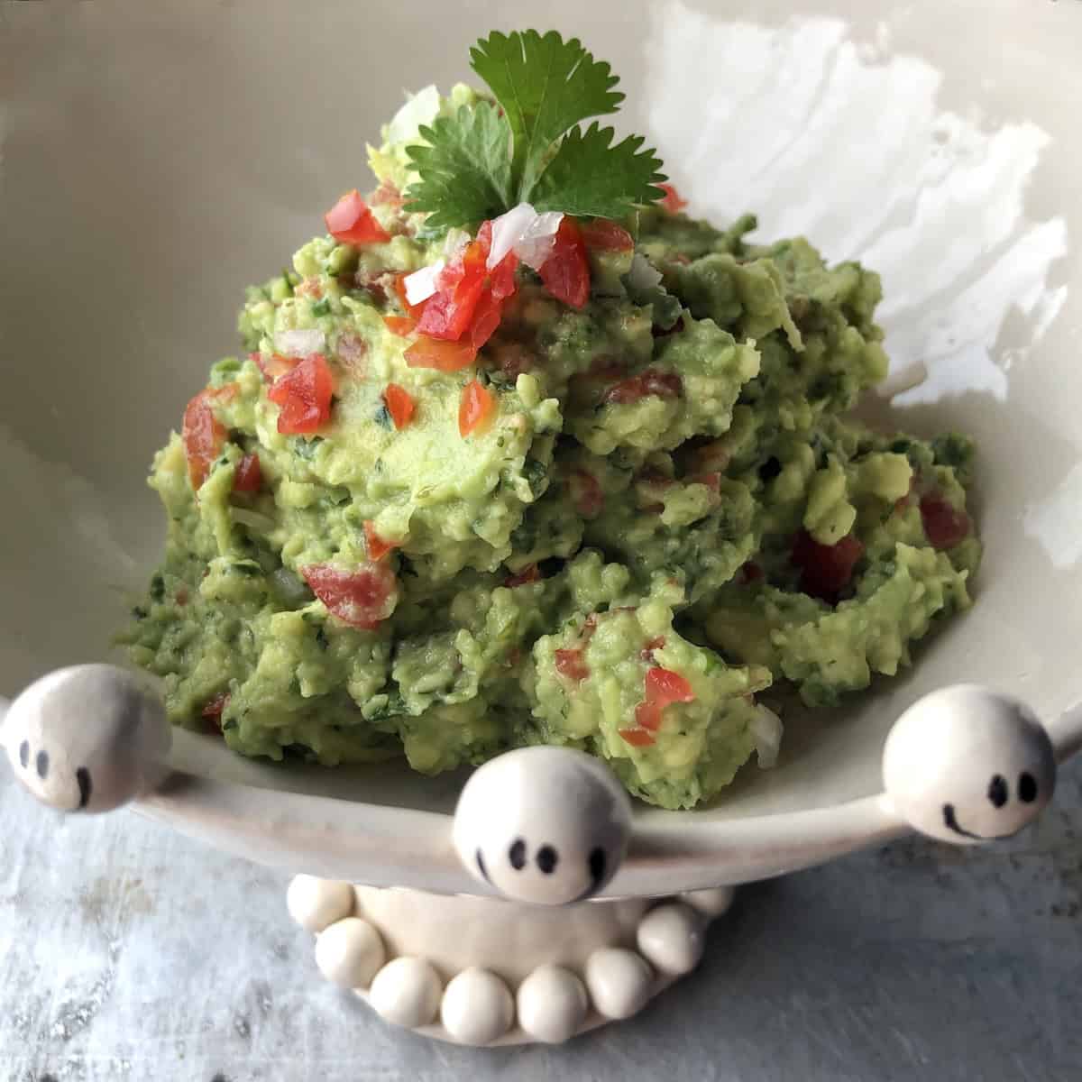 
Mound of homemade guacamole in a white ceramic dish on a pedestal with smiley face ceramic balls surrounding it by  the ceramic artist at The English Boys.
