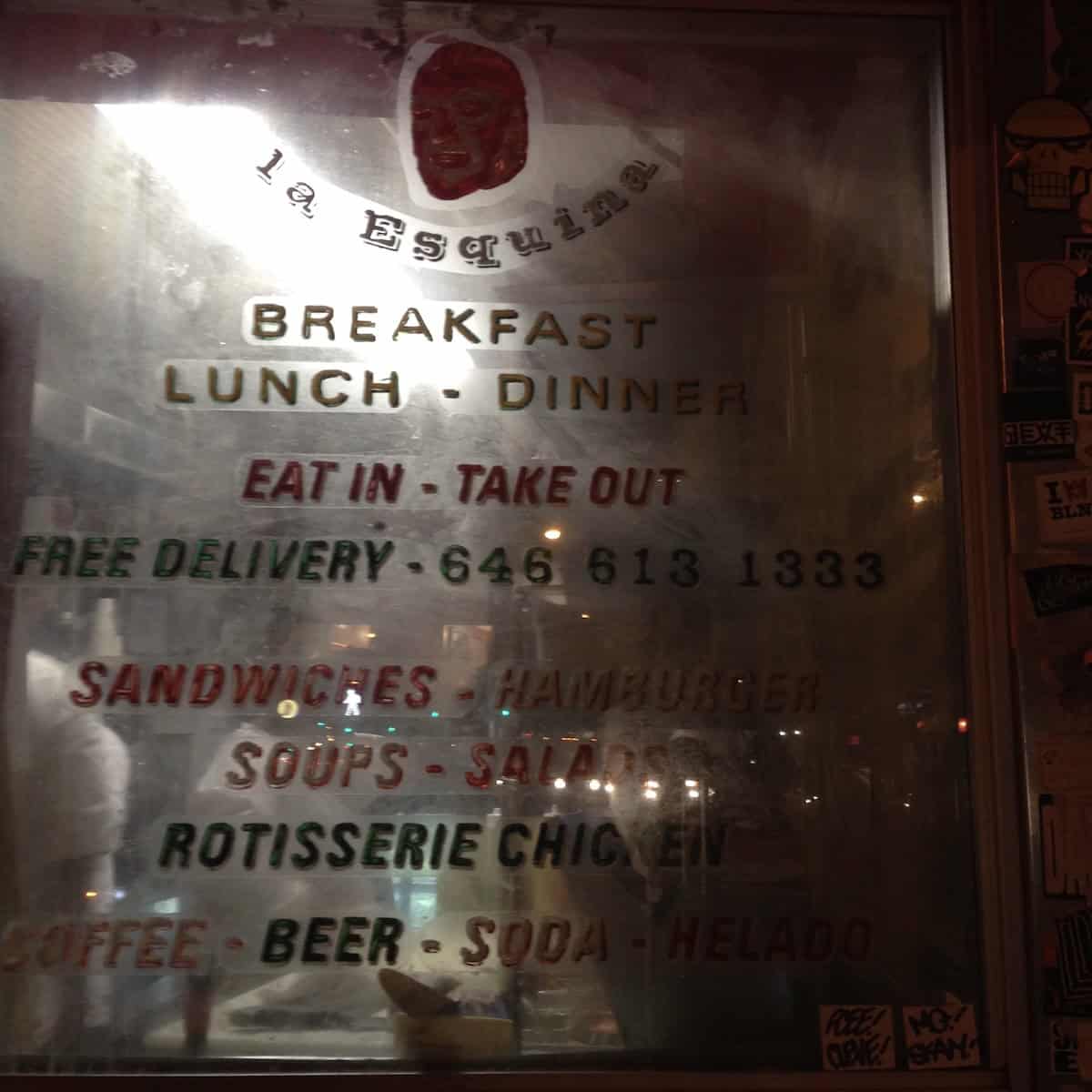 Mirror menu inside La Esquina Mexican restaurant in NYC reading breakfast lunch dinner, eat in take out, free delivery, sandwiches, hamburger, soups, salads, rotisserie chicken, coffee, beer, soda, helado.
