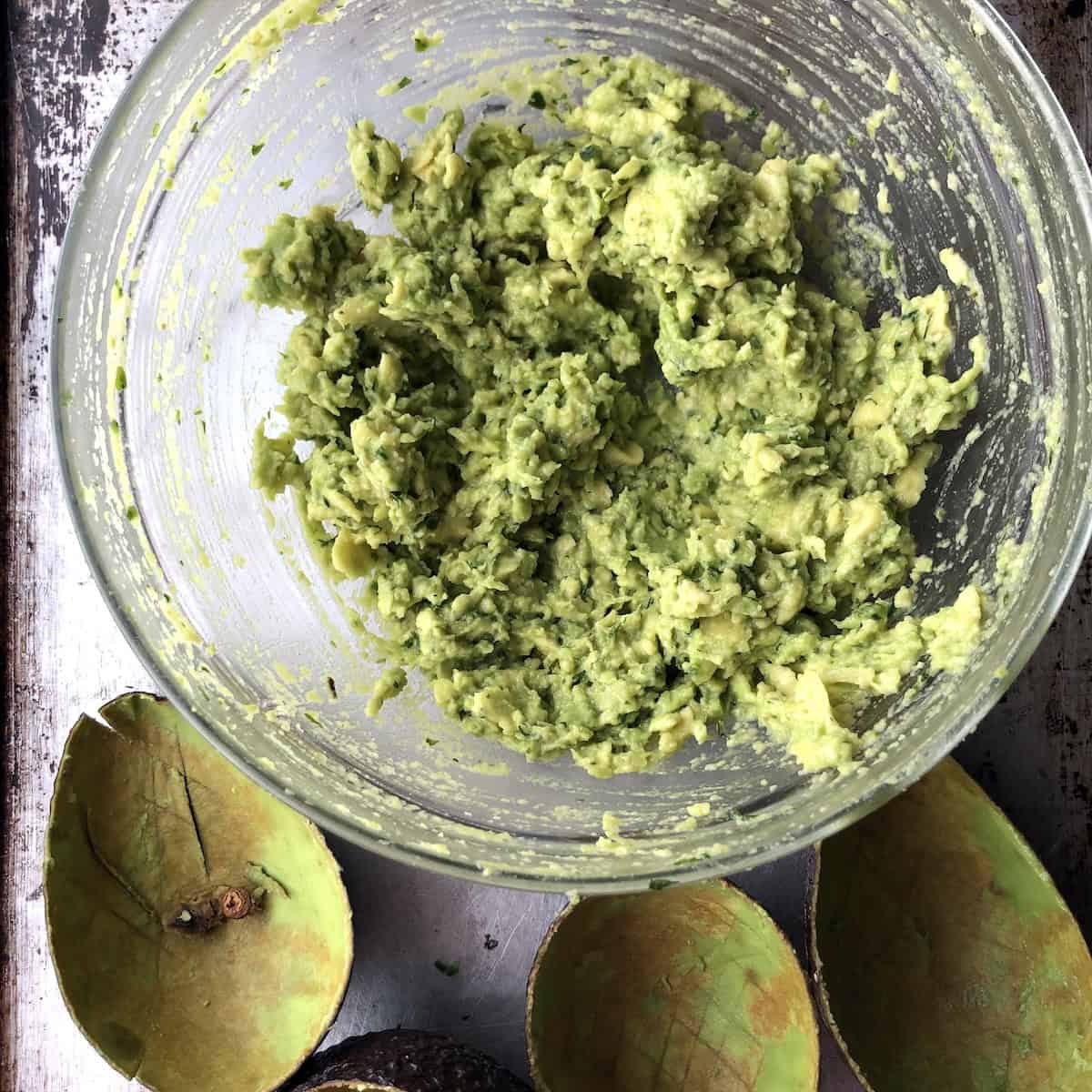 Avocado flesh mashed and mixed with macerated aromatics in a bowl for simple guacamole.