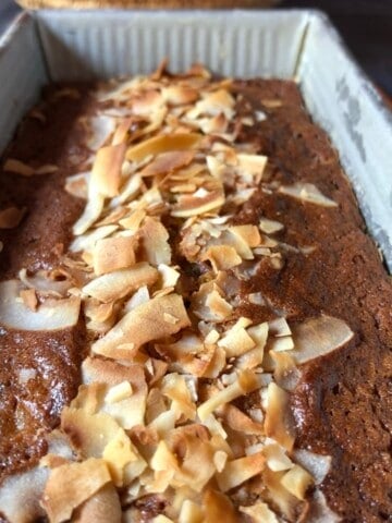 Closeup of a fully baked banana bread with chocolate chips and coconut in a loaf pan with golden brown toasted coconut flakes on top.