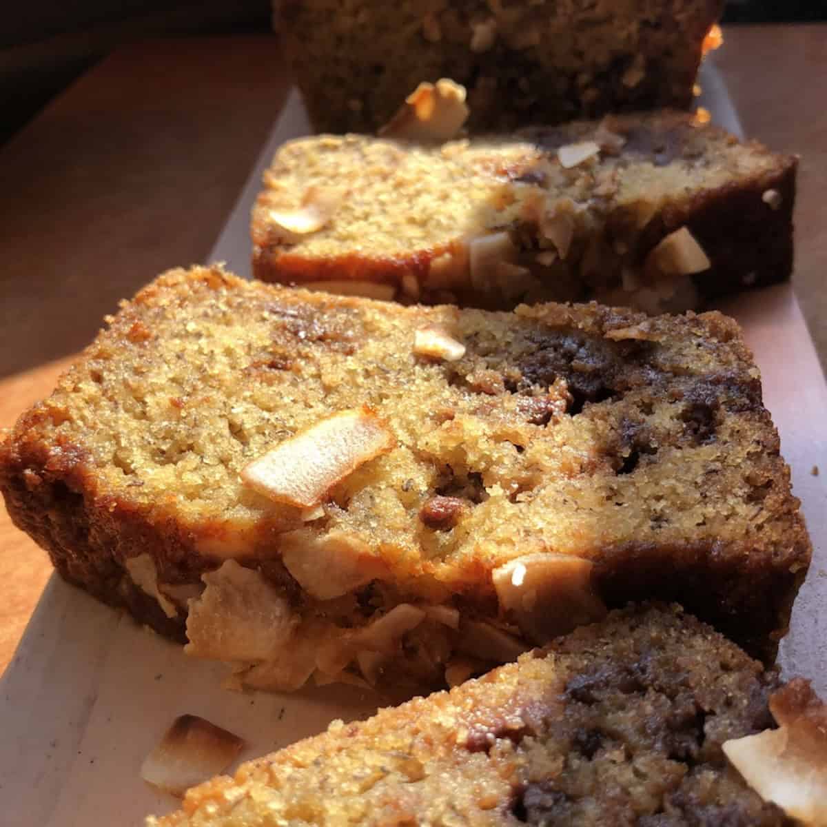 Slices of moist coconut banana bread with chocolate chips on a tray in the sunlight showing the tender crumb with the loaf in the background.