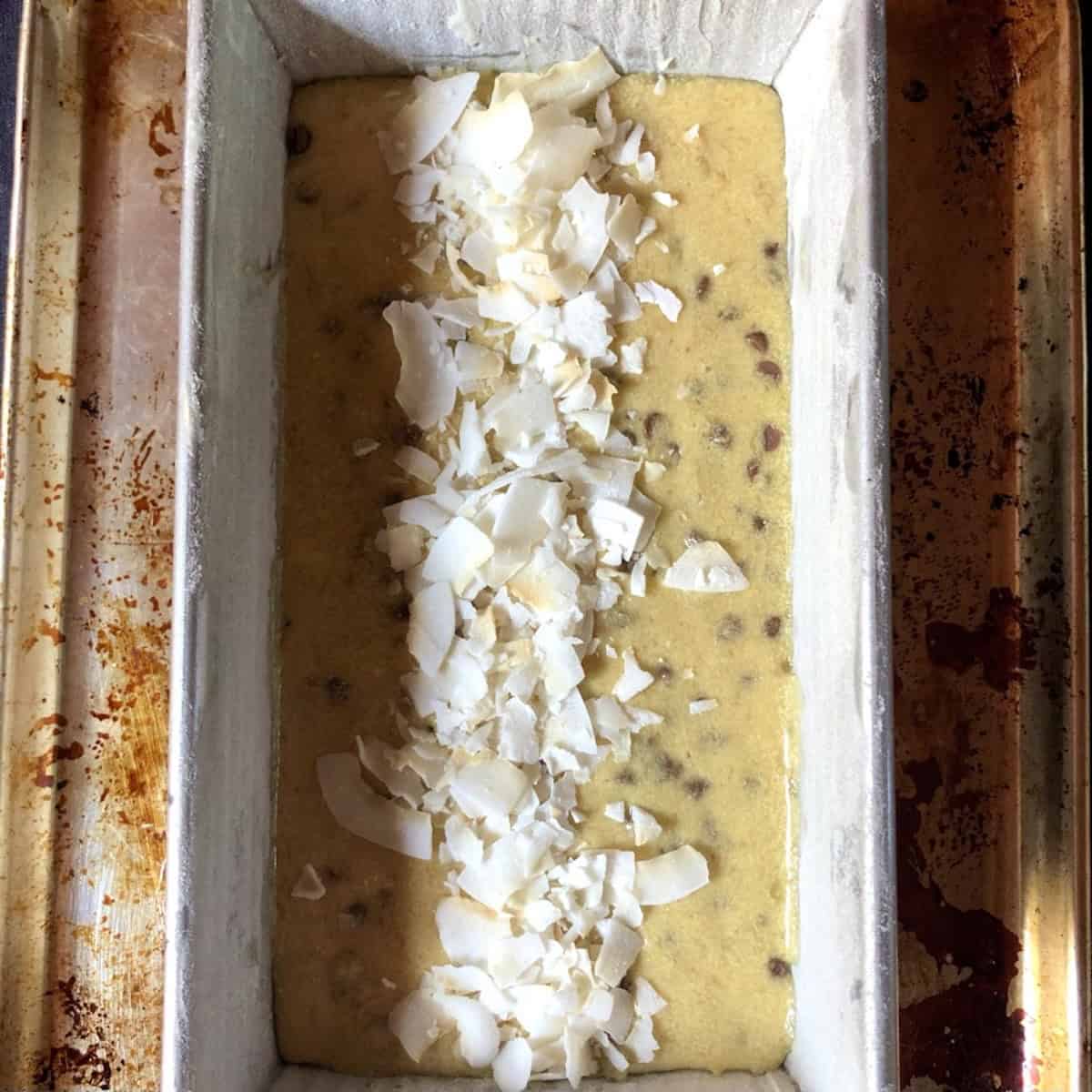 Chocolate coconut banana bread batter in a loaf pan with crispy dehydrated coconut flakes sprinkled down the middle ready to bake.
