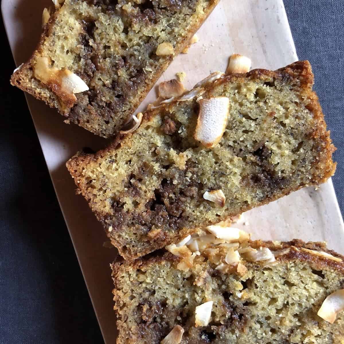 Three slices of banana coconut chocolate chip bread on a serving board showing the moist crumb with melted chocolate chips and coconut throughout.
