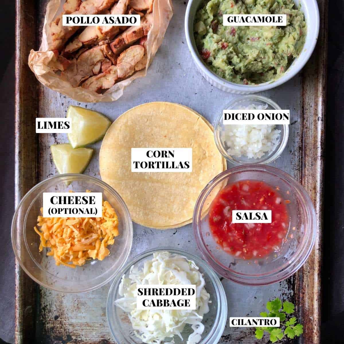 Labeled pollo asado taco ingredients on a cutting board including grilled sliced pollo asado, yellow corn tortillas, finely diced onions, cilantro, homemade fresh salsa, fresh lime wedges, homemade guacamole, and finely shredded white cabbage.
