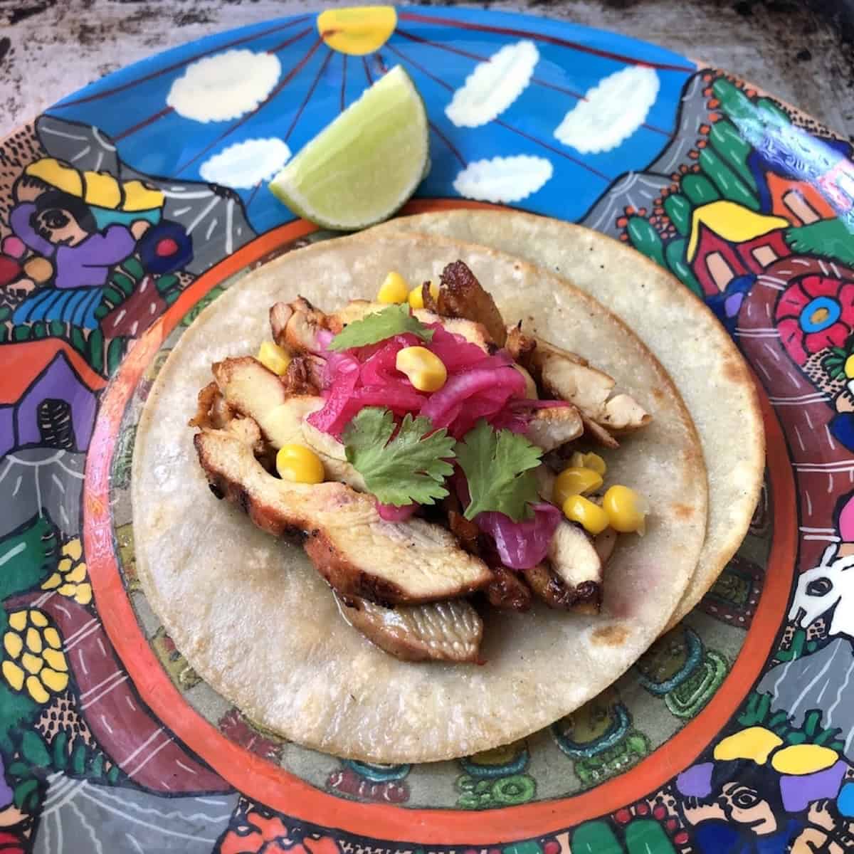 A traditional pollo asado chicken taco garnished with pickled red onions (that are bright pink), cilantro, and sweet corn kernels on two white corn tortillas staggered to hold all the ingredients without breaking.