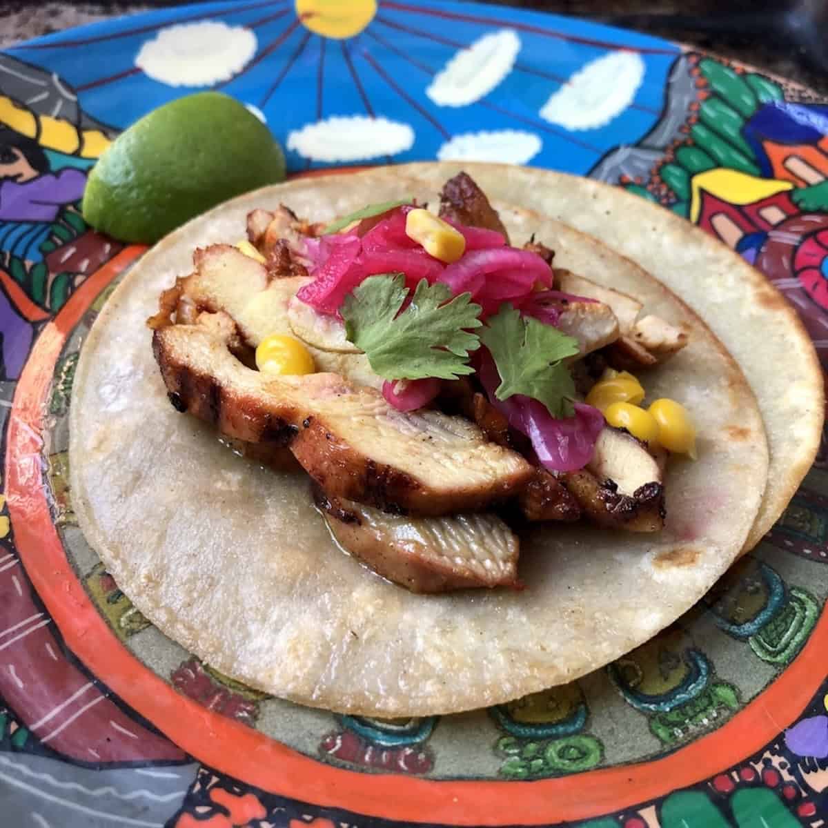 A single Mexican pollo asado chicken taco garnished with pickled red onions, cilantro, and sweet corn kernels on two white corn tortillas staggered to hold all the ingredients without breaking.