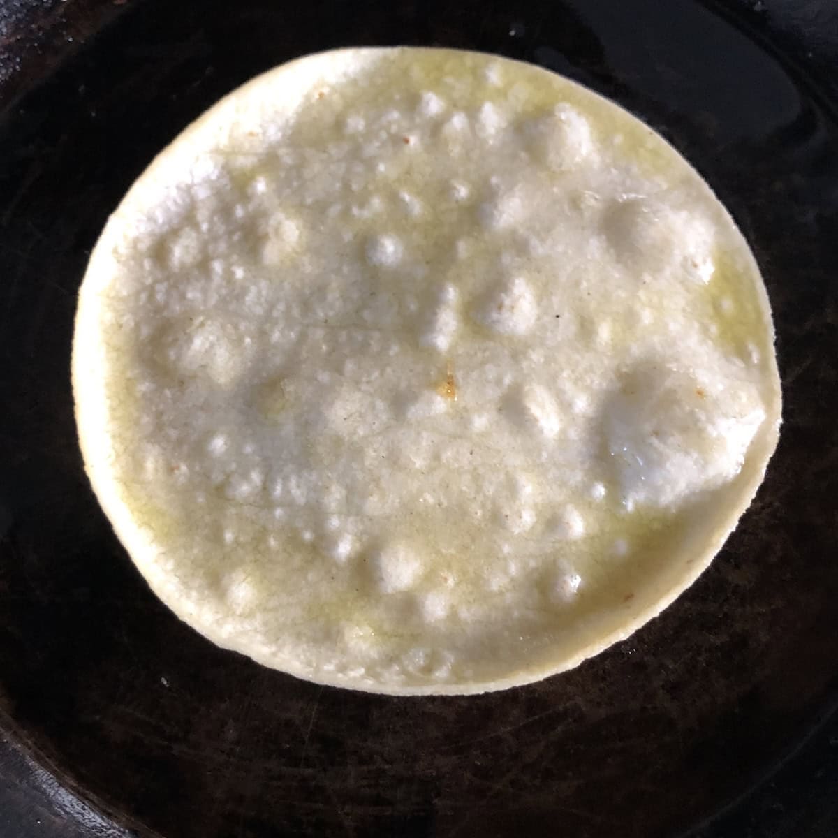 A corn tortilla cooking on the other side until bubbly and bendable after about one minute in the pan.
