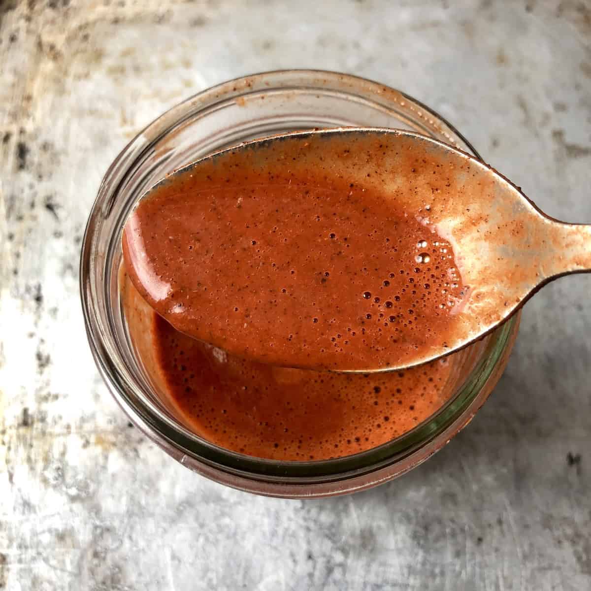 A spoonful of pollo asado marinade held above the jar showing flecks of real spices and the ideal smooth consistency for a homemade pollo asado marinade recipe.