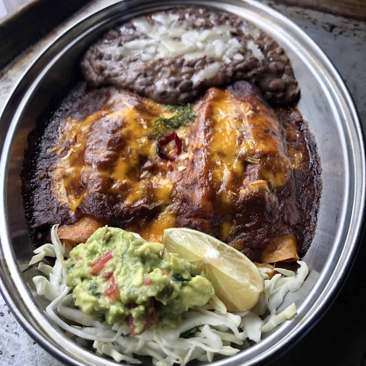 Three pollo asado chicken enchiladas in a stainless steel oval platter with healthy black refried beans, shredded lettuce, homemade guacamole, and a fresh lime wedge showing how to use pollo asado marinade for Mexican recipes.