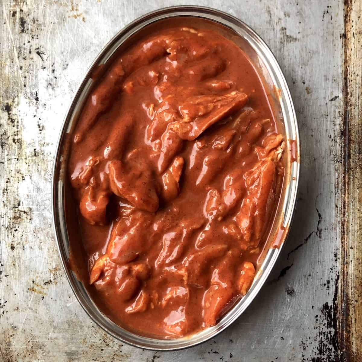 Boneless skinless chicken thighs completely covered in bright red-orange pollo asado marinade on both sides in a stainless steel oval dish ready to cover, refrigerate, and marinate for 12-14 hours (showing how to marinate chicken for pollo asado).
