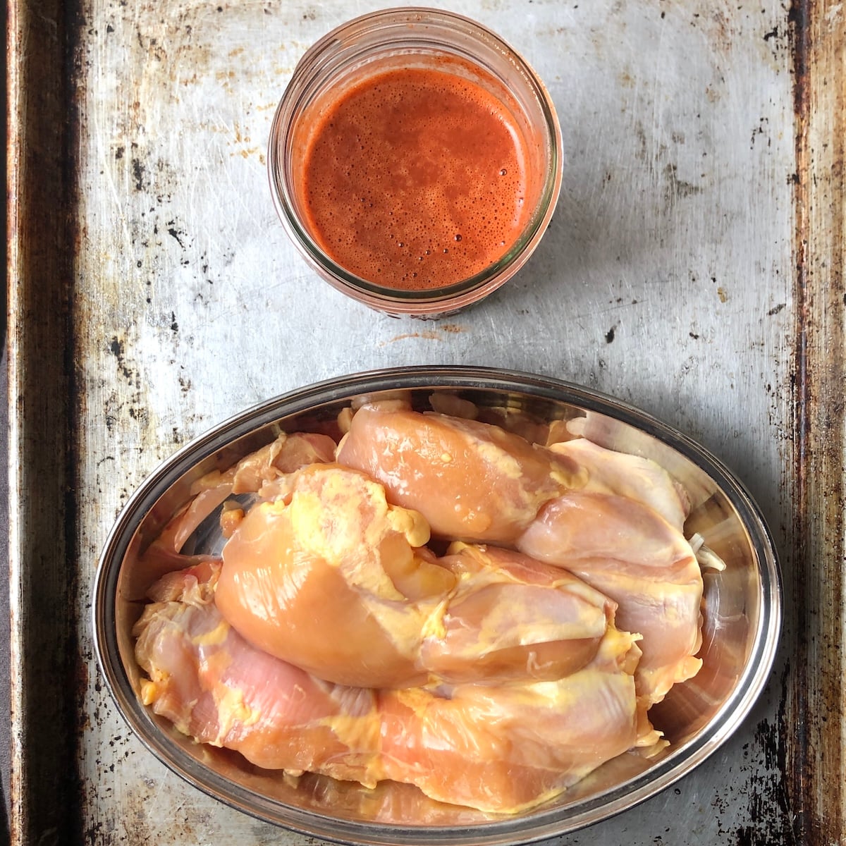 Boneless skinless chicken thighs in a stainless steel oval container next to a jar of pollo asado mexican chicken marinade ready to marinate.