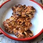 Two grilled pollo asado chicken thighs with char marks and juices in a red-rimmed serving bowl resting before slicing, pollo asado recipe.