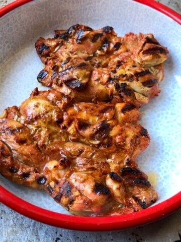 Two grilled pollo asado chicken thighs with char marks and juices in a red-rimmed serving bowl resting before slicing, pollo asado recipe.