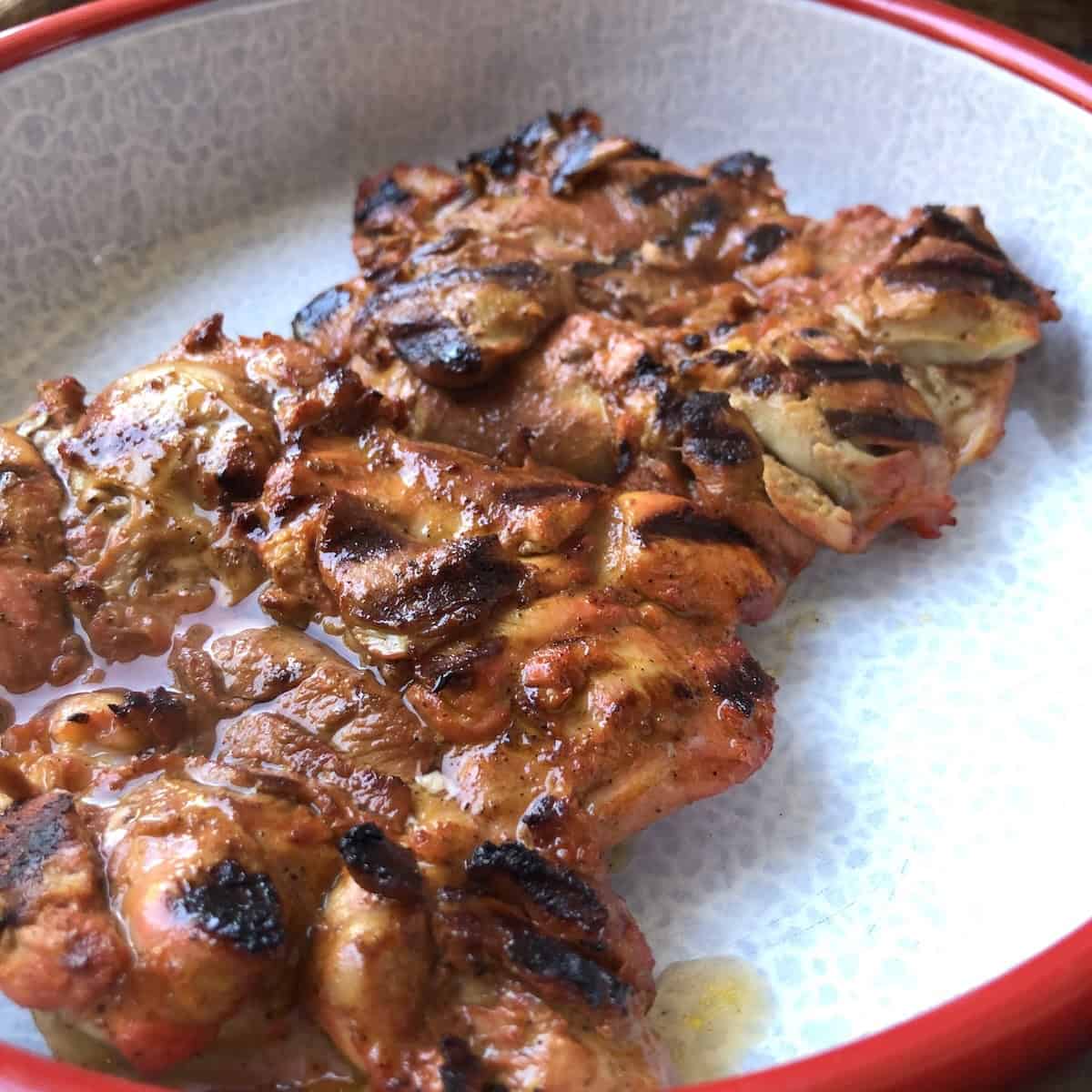 Two grilled pollo asado chicken thighs with char marks and juices in a red-rimmed serving bowl resting before slicing, pollo asado recipe.
