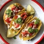 Three pollo asado tacos in a taco holder on a serving platter showing how to present chicken tacos al carbon garnished with onions, clilantro, fresh salsa, guacamole and shreeded white cabbage with 2 lime wedges.