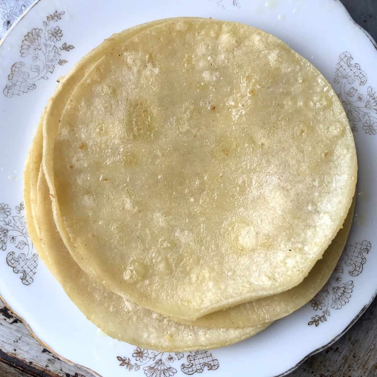 A stack of lightly cooked warmed corn tortillas on a plate ready to use for pollo asado tacos.