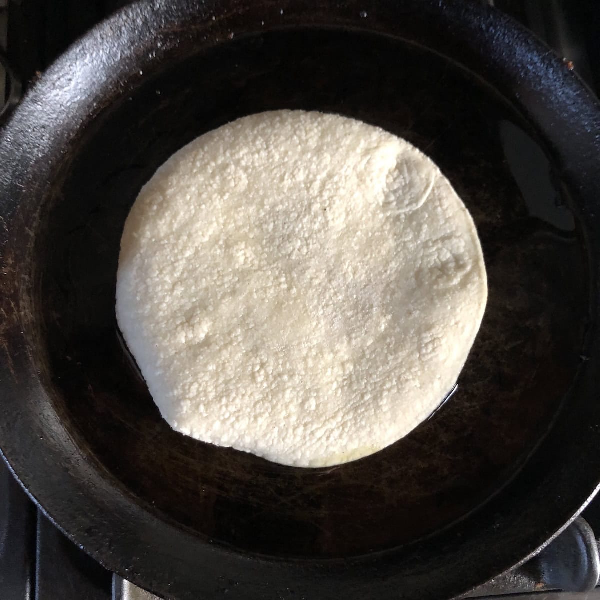 A dry corn tortilla added to a lightly oiled carbon steel crepe pan to warm before using for homemade chicken tacos.