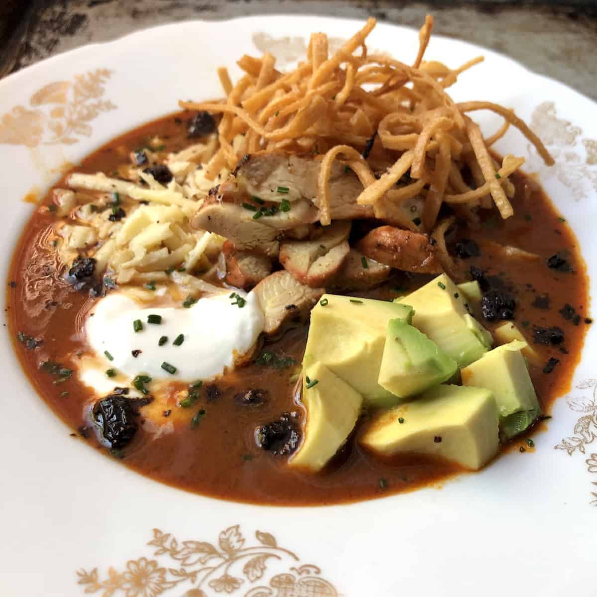 Authentic chicken tortilla soup with crispy fried tortilla strips, sliced pollo asado, diced avocado, crema, monterey jack cheese, fried mulato chile, and chives in a deep red broth.