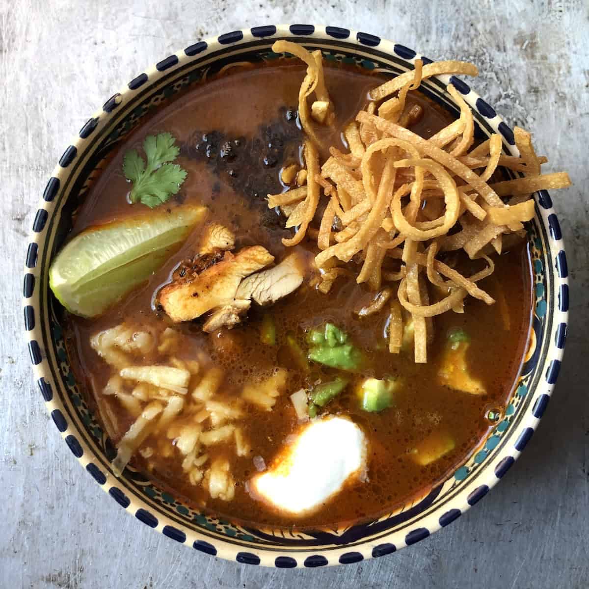 Top down view of a bowl of sopa de tortilla con pollo with fried tortilla strips, pollo asado, corn, cheese, avocado, crema, and fried mulato chile and one leaf of cilantro.
