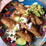 Three crispy golden brown chicken flautas on a colorful Mexican platter on shredded lettuce topped with salsa fresca, sour cream, jalapeno slices, guacamole, and a lime wedge.