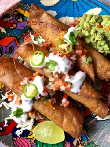 Three crispy golden brown chicken flautas on a colorful Mexican platter on shredded lettuce topped with salsa fresca, sour cream, jalapeno slices, guacamole, and a lime wedge.