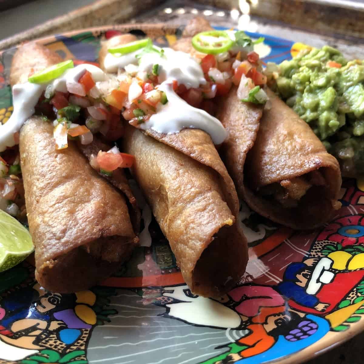 Three crispy chicken flautas on a colorful Mexican platter on shredded lettuce topped with salsa fresca, sour cream, jalapeno slices, guacamole, and a lime wedge side view. 
