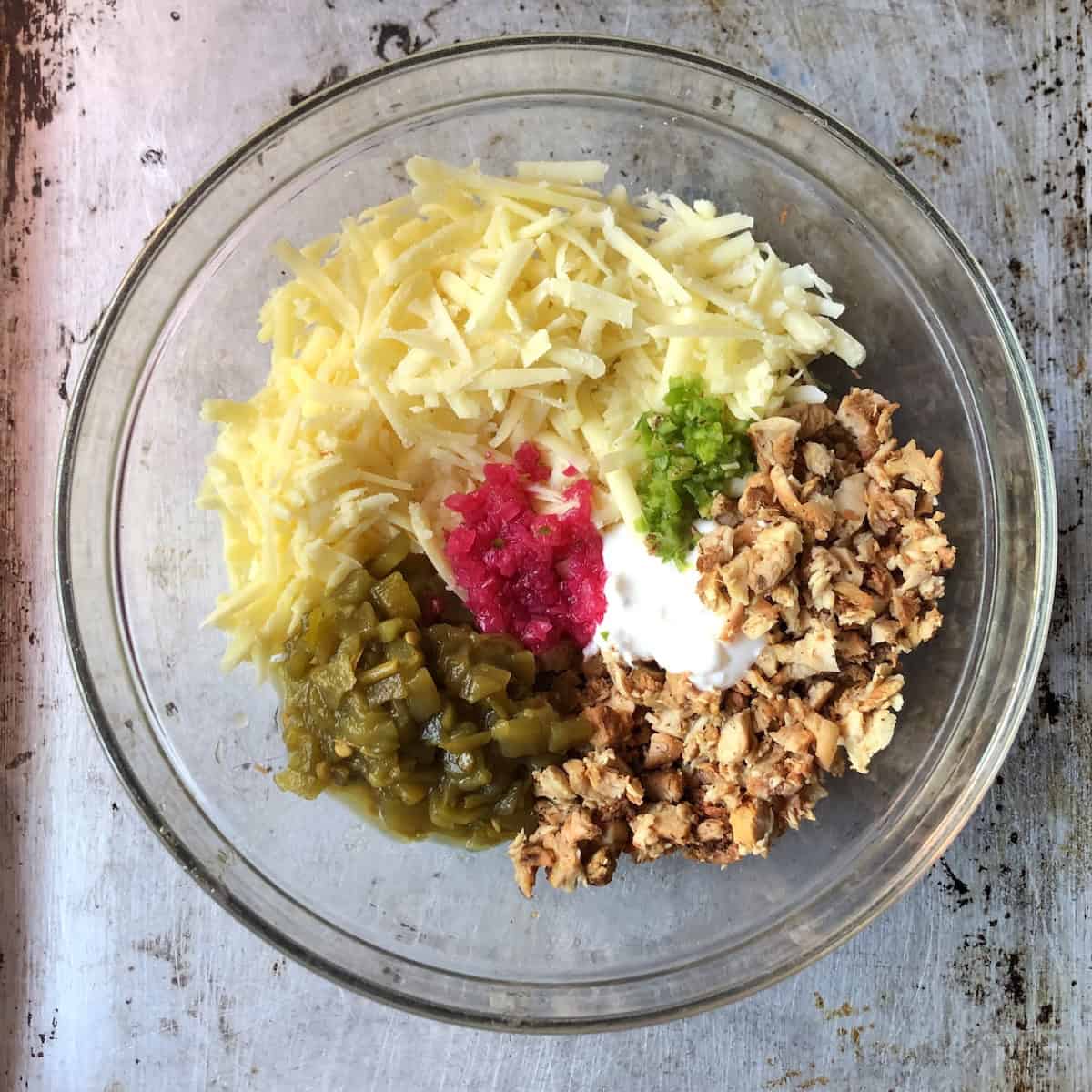 Chicken flauta filling ingredients in a mixing bowl including diced pollo asado, shredded white cheddar, green chiles, fresh jalapeno, sour cream, and pickled red onions.
