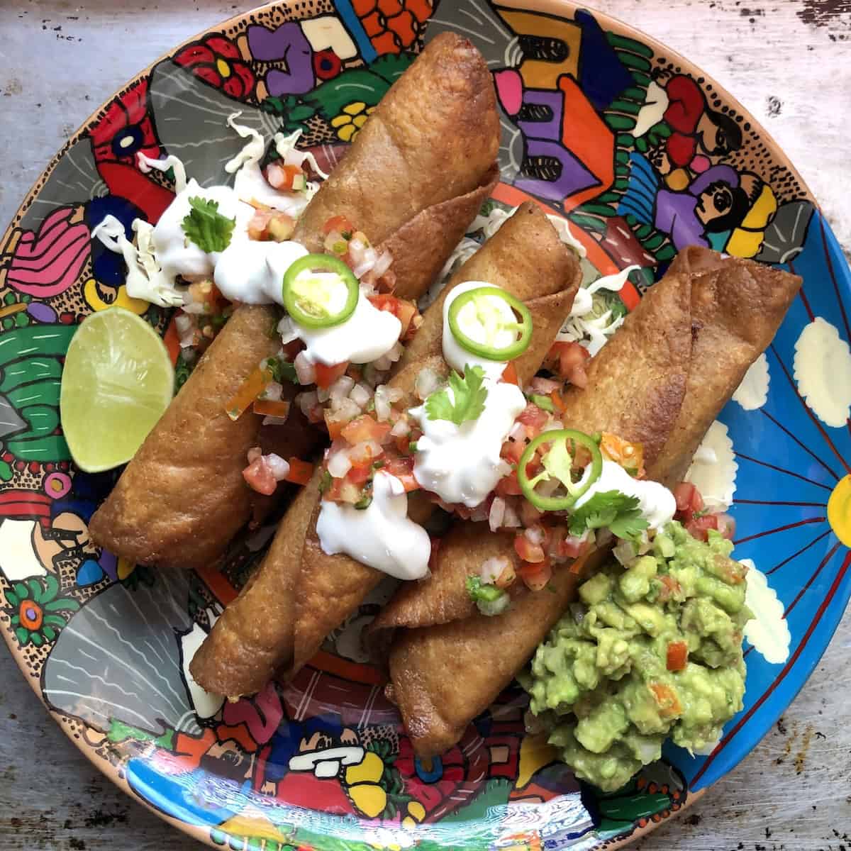Three crispy flautas mexicanas on a colorful platter with shredded lettuce, homemade guacamole, salsa fresca, sour cream, fresh jalapeno slices, and a lime wedge.
