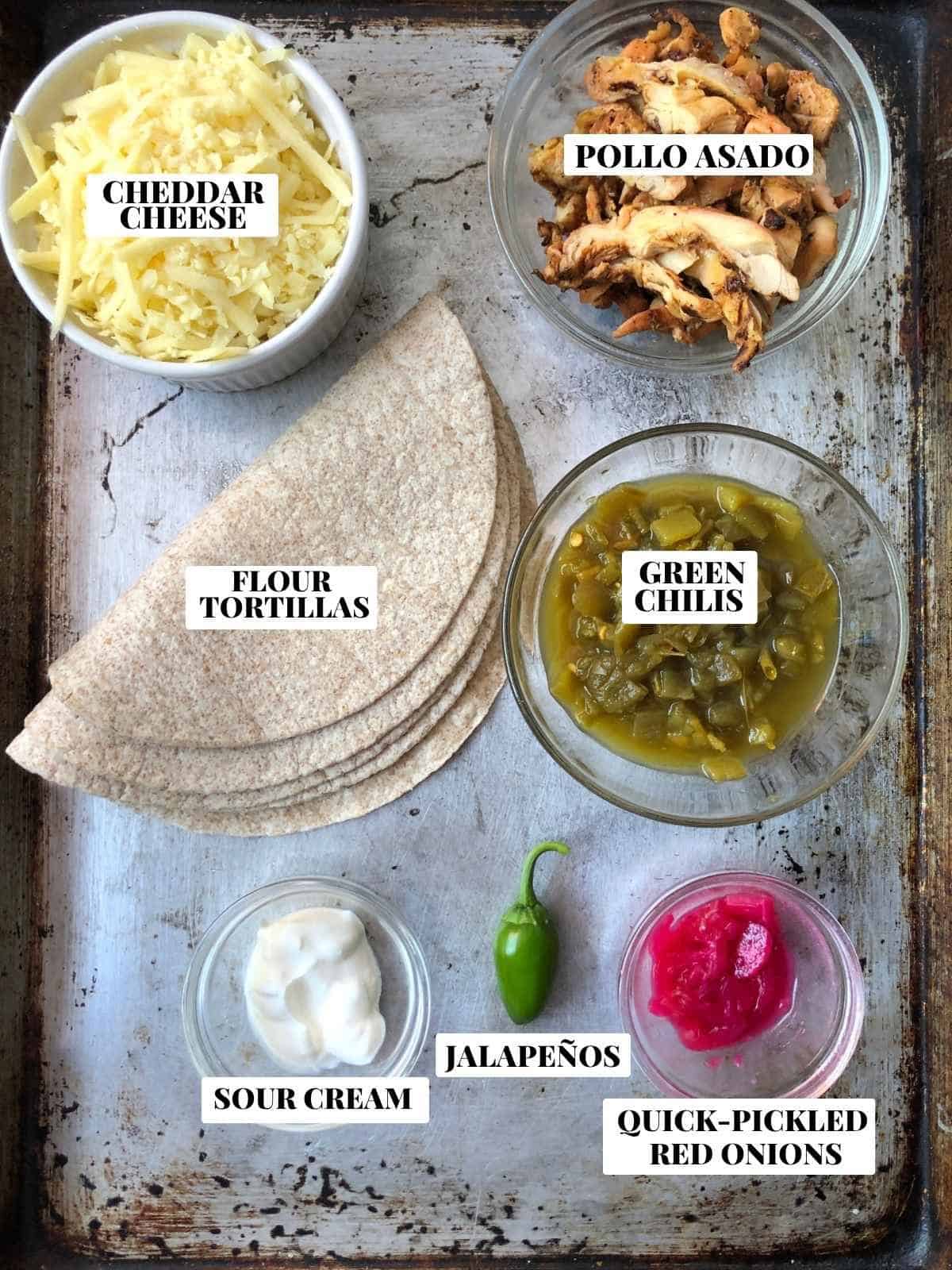 Labeled chicken flautas recipe ingredients on a sheet tray including pollo asado chicken thigh meat, whole wheat flour tortillas, white cheddar cheese shredded, sour cream, green chilis, quick pickled red onions, and jalapeno
