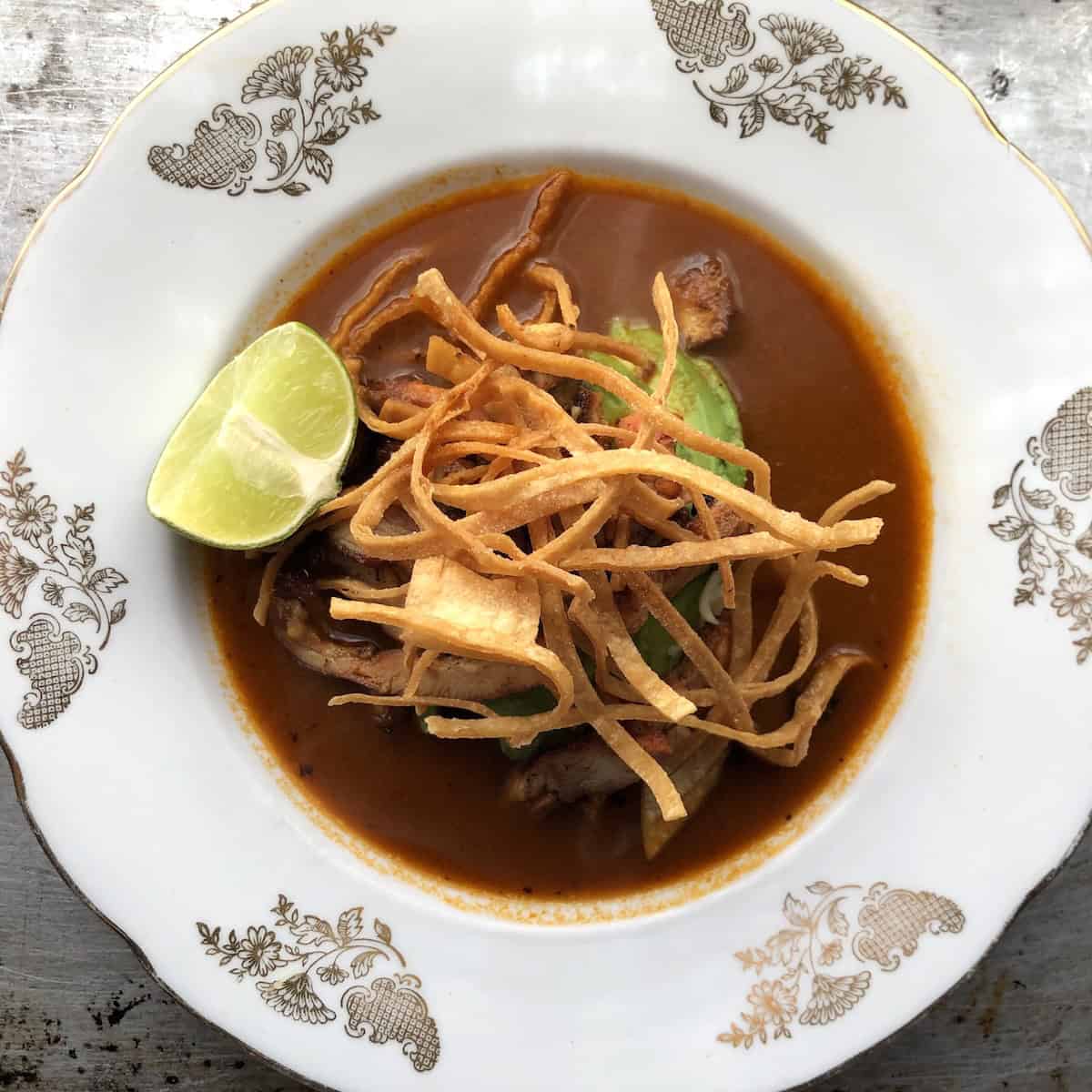 Top down view of a bowl of chicken lime tortilla soup with crispy tortilla strips, sliced chicken, crema, cheese, avocado, and a lime wedge in a rich chile-tomato broth.


