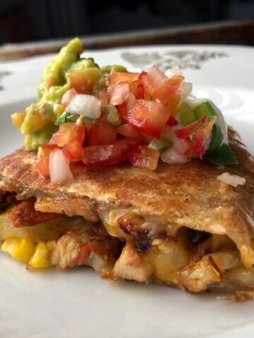 A golden brown chicken quesadilla wedge on a plate topped with guacamole, salsa fresca, and sour cream with melted cheese, chicken, and corn filling visible, chicken quesadilla recipe.