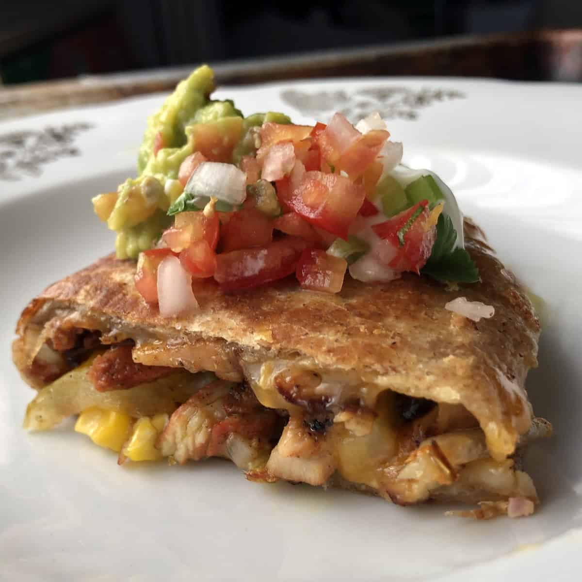 A golden brown chicken quesadilla wedge on a plate topped with guacamole, salsa fresca, and sour cream with melted cheese, chicken, and corn filling visible, chicken quesadilla recipe.
