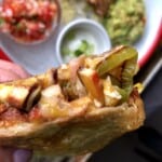 Hand holding a chicken quesadilla slice showing melted cheese, pollo asado chicken, caramelized peppers, and onions with salsa, sour cream, and guacamole in the background.