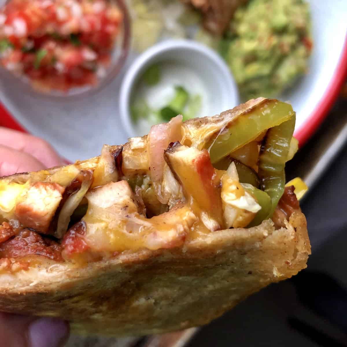 Hand holding a chicken quesadilla slice showing melted cheese, pollo asado chicken, caramelized peppers, and onions with salsa, sour cream, and guacamole in the background.
