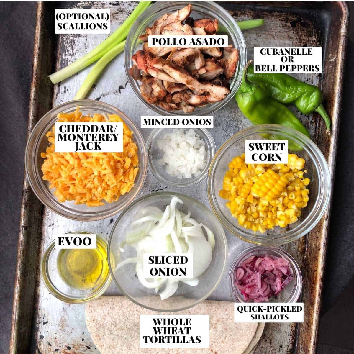 Labeled chicken quesadilla ingredients on a baking tray including sliced pollo asado chicken, whole wheat flour tortillas, cubanelle peppers, sliced and minced onions, corn, cheddar and monterey jack cheese, olive oil, quick pickled shallots, and scallions.
