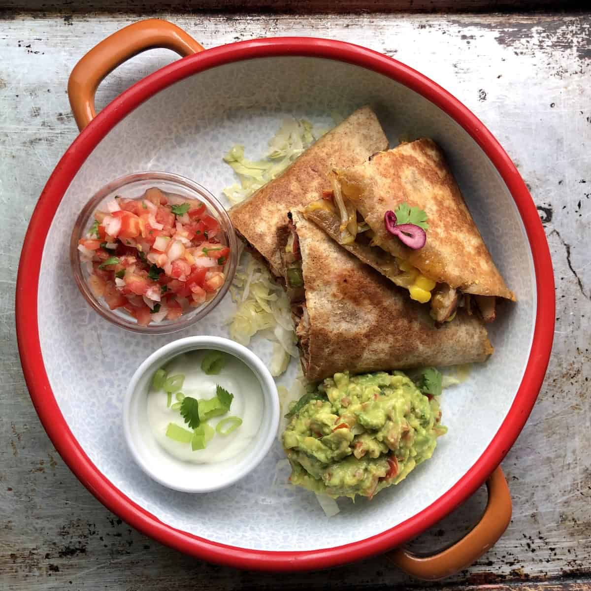 Sliced chicken quesadilla on a serving platter with homemade salsa fresca, sour cream, and guacamole on a bed of shredded iceberg lettuce with melty fillings in full view.