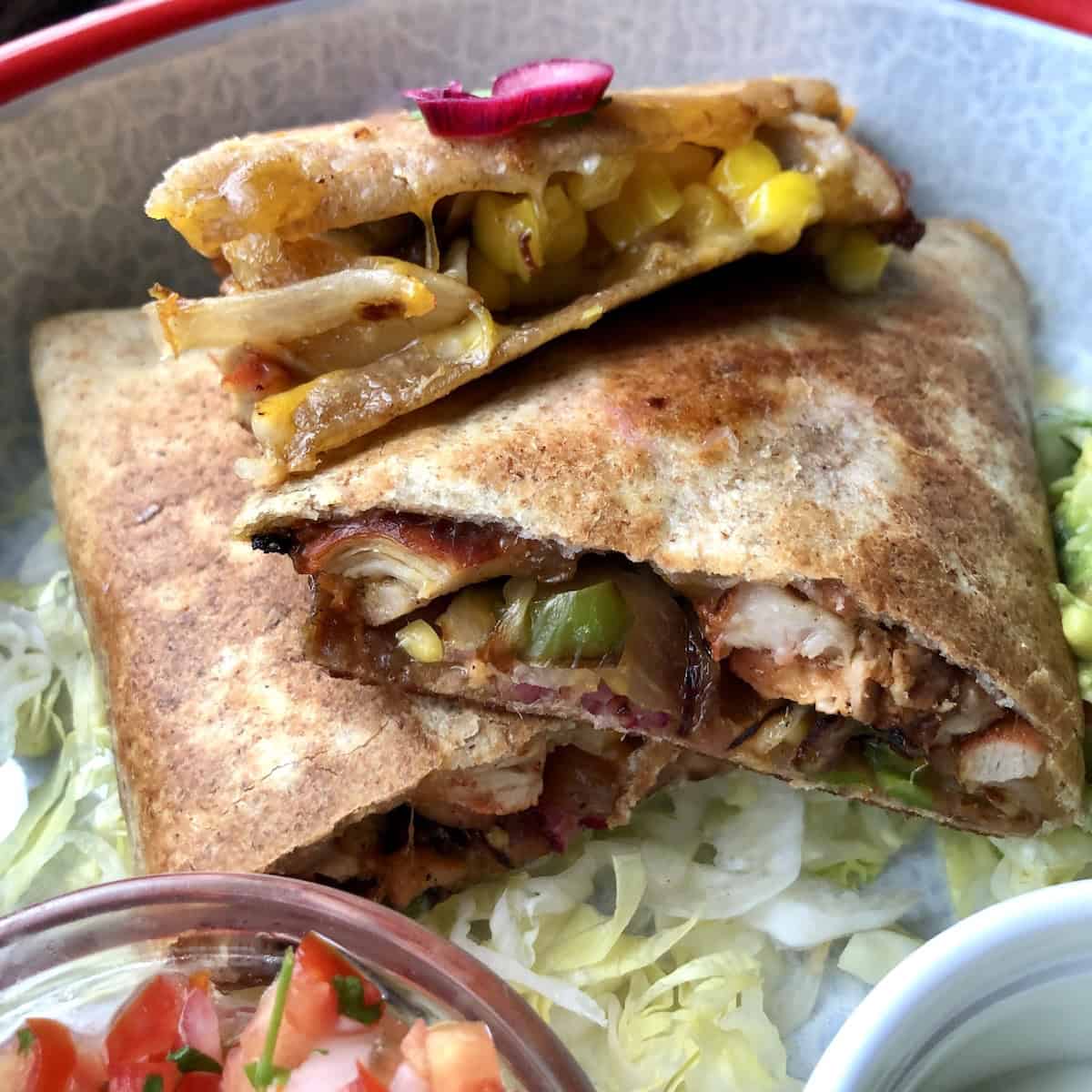 Sliced chicken quesadillas stacked in a serving platter with toppings alongside and pink pickled red onion and cilantro on top.
