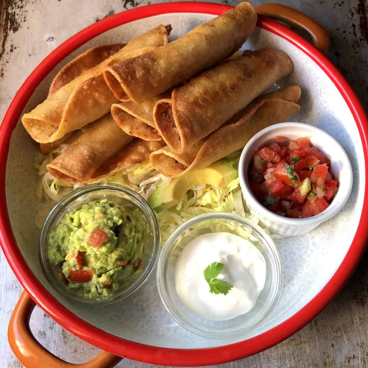 Crispy chicken taquitos recipe piled high on a serving platter on shredded lettuce with salsa fresca, sour cream, guacamole, and avocado slices on the side.

