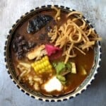 A bowl of chicken tortilla soup with fried tortilla strips, pollo asado, black beans, pickled red onion, corn, monterey jack cheese, avocado, crema, and fried mulato chile in a deep red broth.