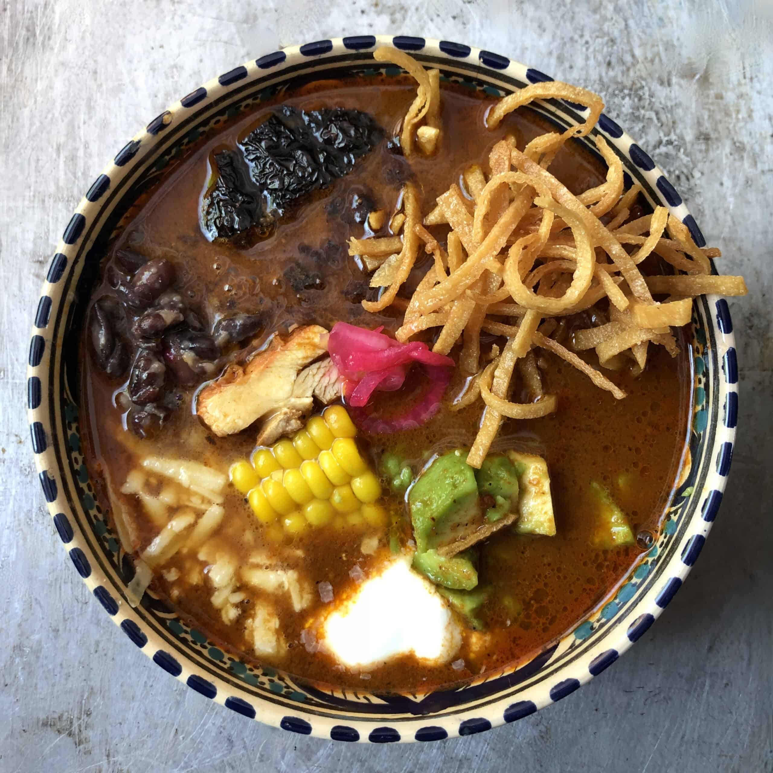 A bowl of chicken tortilla soup with fried tortilla strips, pollo asado, black beans, pickled red onion, corn, monterey jack cheese, avocado, crema, and fried mulato chile in a deep red broth.
