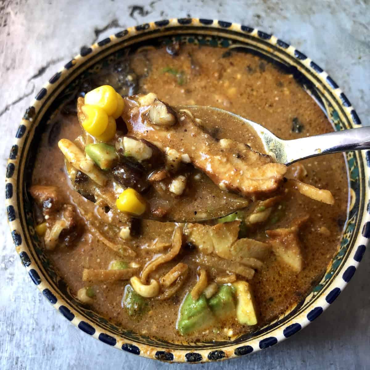 A spoonful of creamy chicken tortilla soup showing corn, avocado, black beans, tortilla strips, and chicken.
